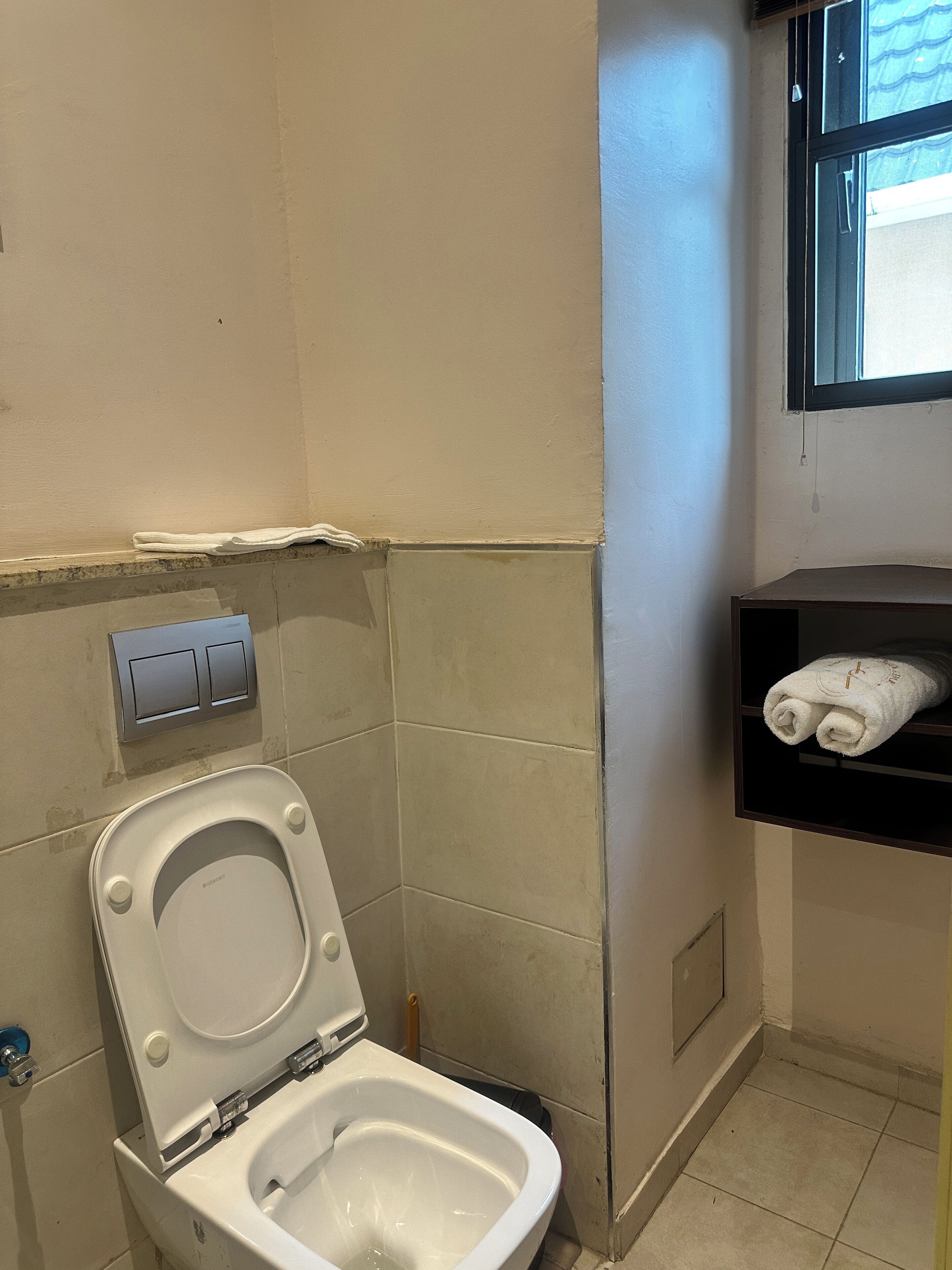 Un baño con inodoro blanco, un perchero con toallas y una ventana con persianas.
