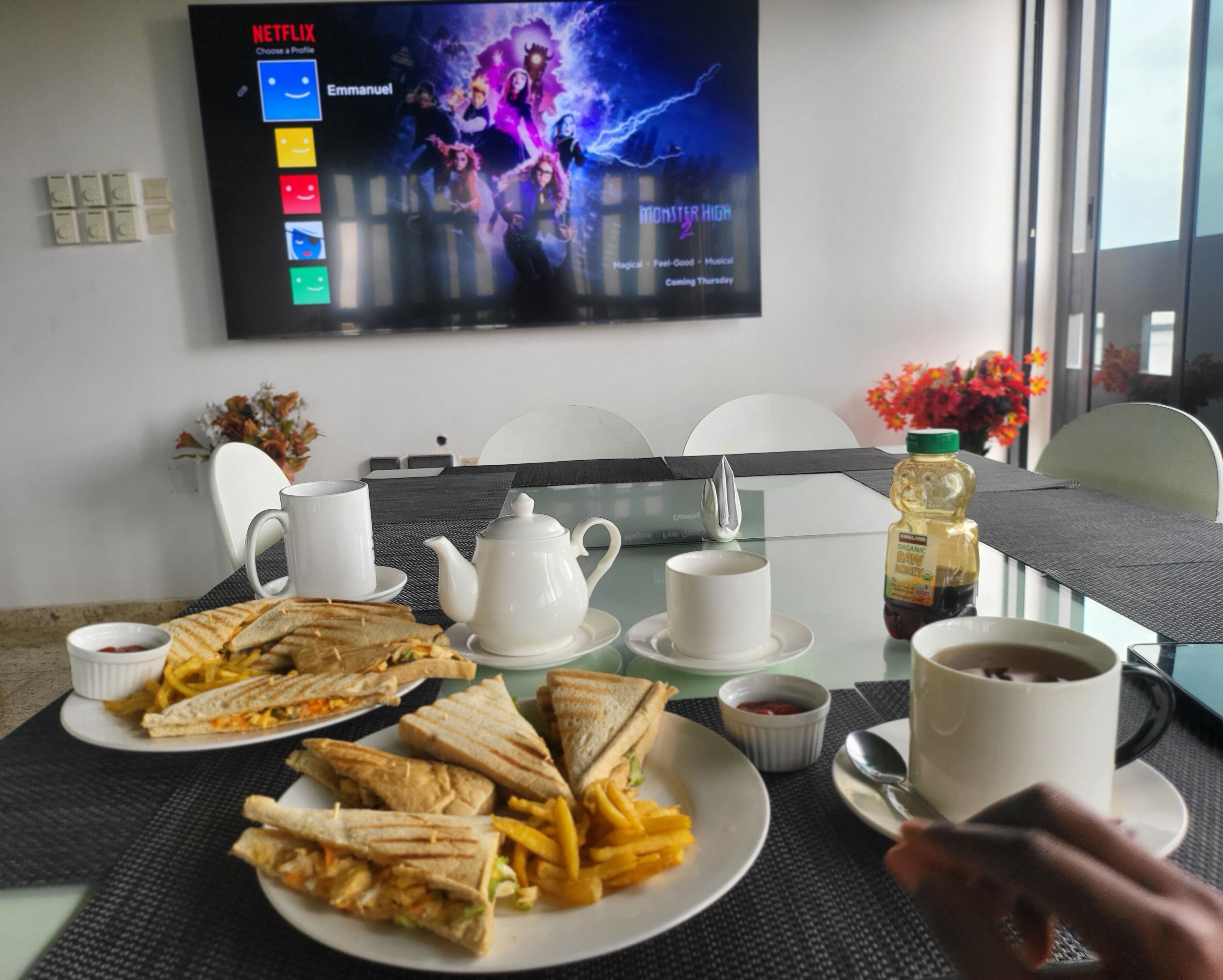A dining table with sandwiches, fries, and a bottle of oil, a teapot, and cups, with a TV displaying a movie poster in the background.