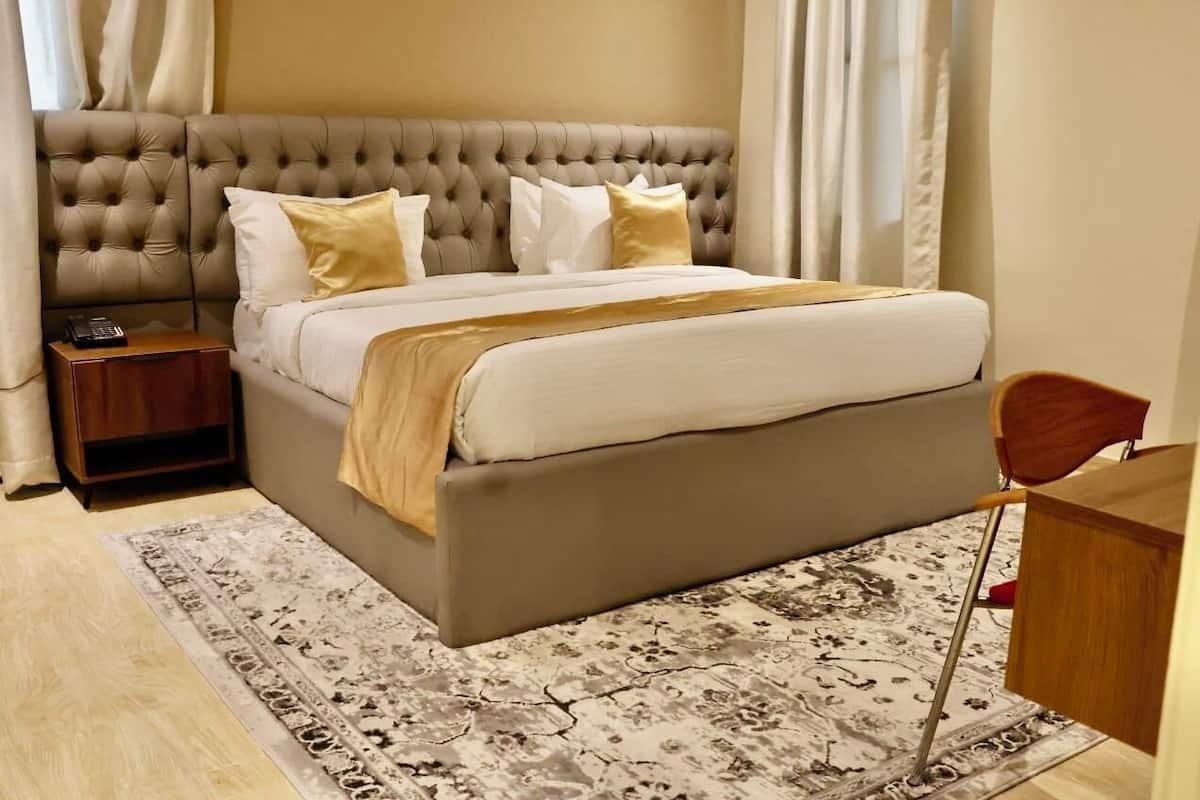 Un dormitorio con cabecera capitonada, cama con ropa de cama blanca y beige, mesita de noche con un teléfono y una alfombra estampada.