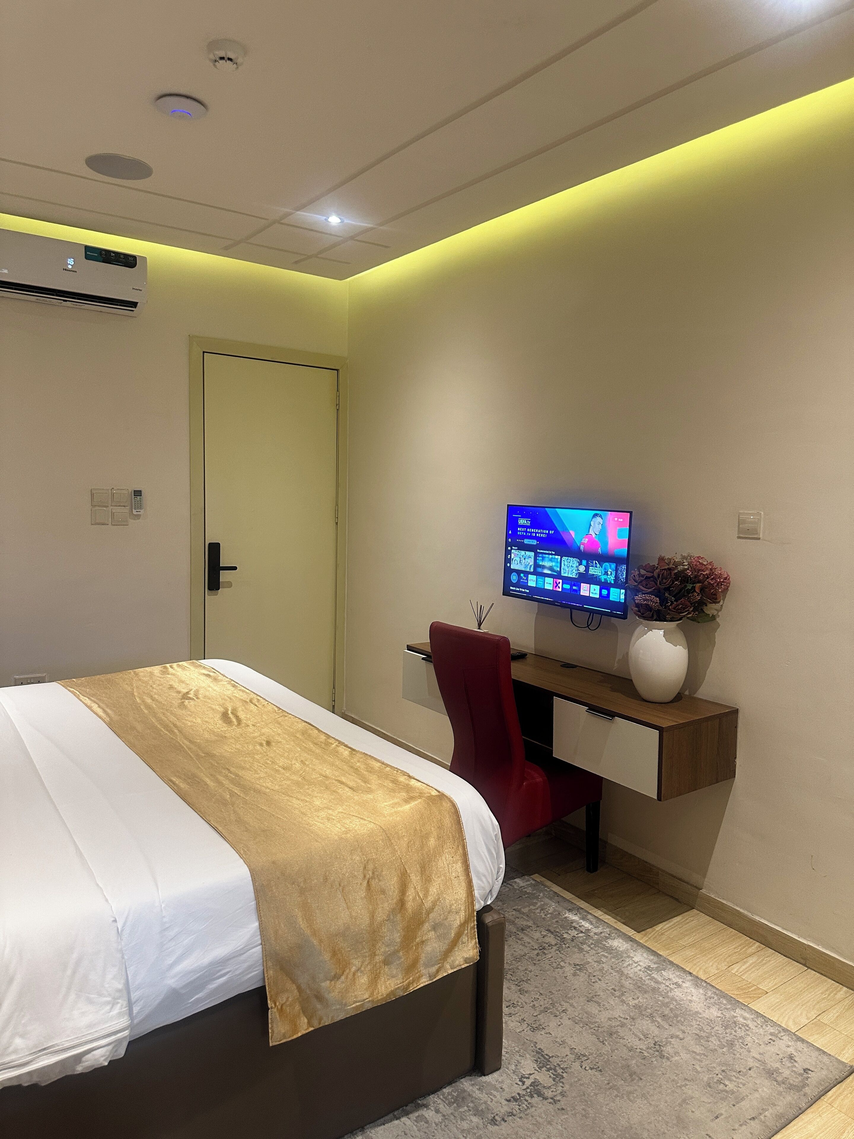 A modern hotel room with a bed, a desk, a red chair, a TV, and a vase with flowers.