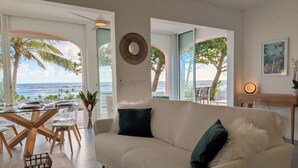 Ein Wohnzimmer mit einer Couch, einem Couchtisch und einem Esstisch für zwei Personen. Durch große Fenster ist der Blick auf den Ozean.