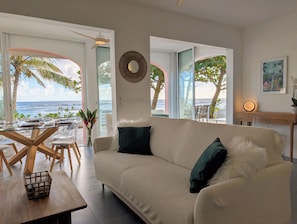 Ein Wohnzimmer mit einer Couch, einem Couchtisch und einem Esstisch für zwei Personen. Durch große Fenster ist der Blick auf den Ozean.
