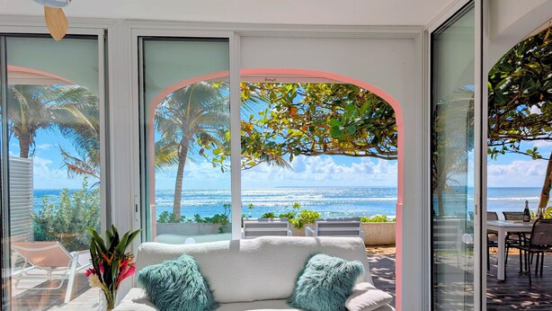 Ein modernes Wohnzimmer mit einem weißen Sofa, zwei türkisfarbenen Kissen und einem kleinen Holztisch. Eine große Glasschiebetür führt in einen Außenbereich mit Blick auf das Meer und Palmen.