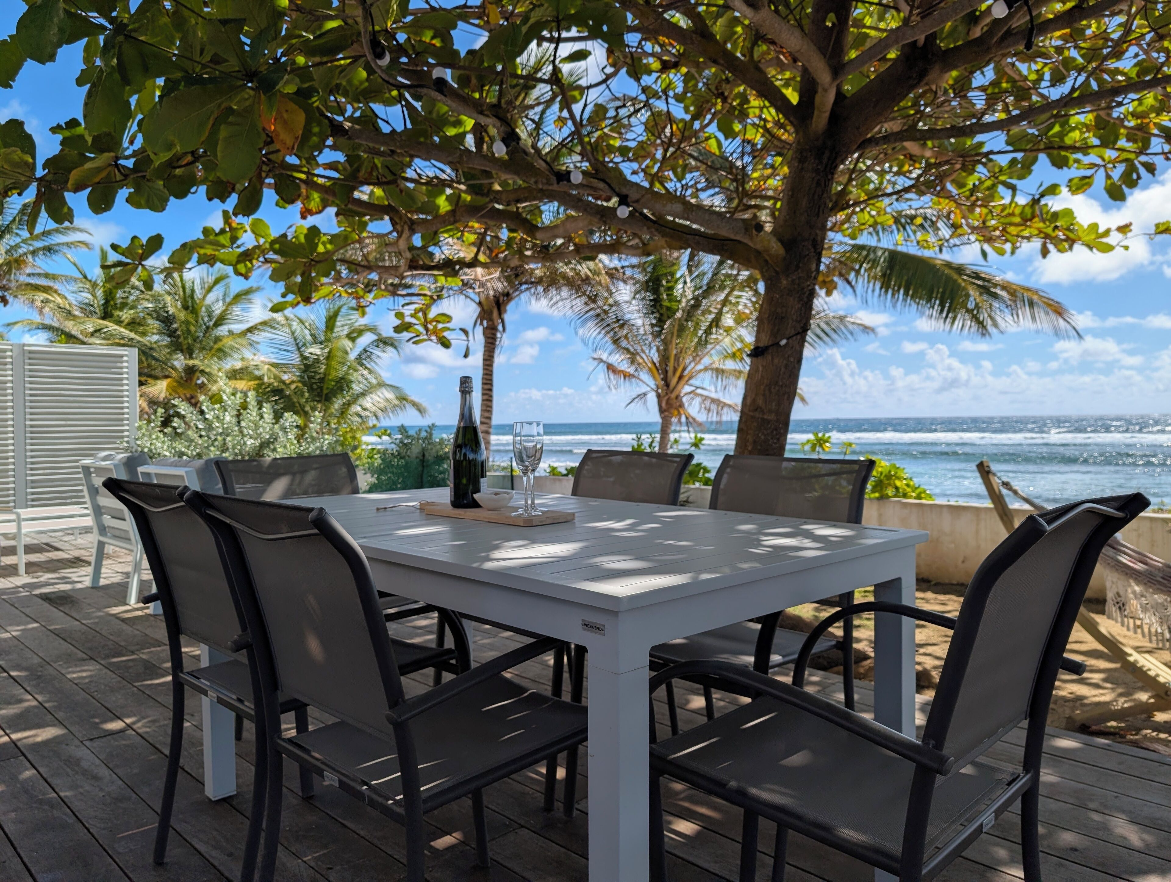 Ein Tisch im Freien mit Stühlen, einer Flasche und einem Glas, mit Blick auf einen Strand mit Palmen und dem Meer.
