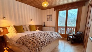 Ein Schlafzimmer mit Holzbalkendecke, einem großen Bett mit floralem Bettzeug, einem Nachttisch, einem Sessel und einem Fenster mit Bergblick.