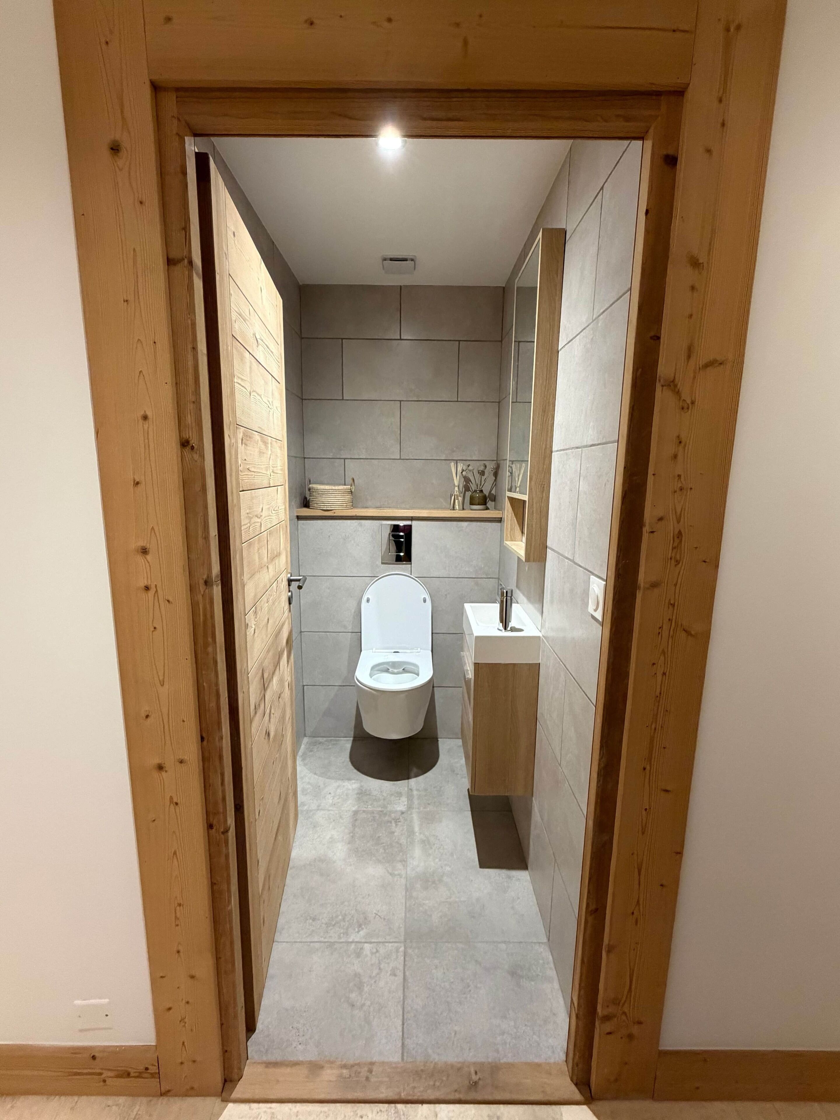 A narrow, wooden-framed bathroom with a toilet, sink, and tiled walls.