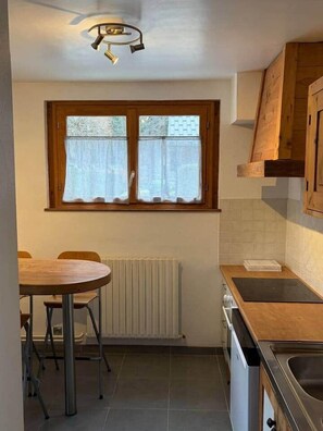Une cuisine de taille réduite comprenant une table à manger, des chaises, une fenêtre avec des rideaux, un radiateur et une cuisinière.