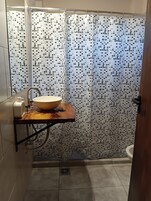 Un baño con piso de baldosas, encimera de madera para el lavamanos, un lavabo blanco, un cabezal de ducha manual y cortina de ducha con diseño de mosaico.