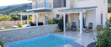 A two-story house with a swimming pool, outdoor seating, and a view of the hills.