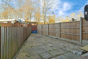 Een betegelde achtertuin met een houten schutting, een bakstenen constructie en bladerloze bomen op de achtergrond.