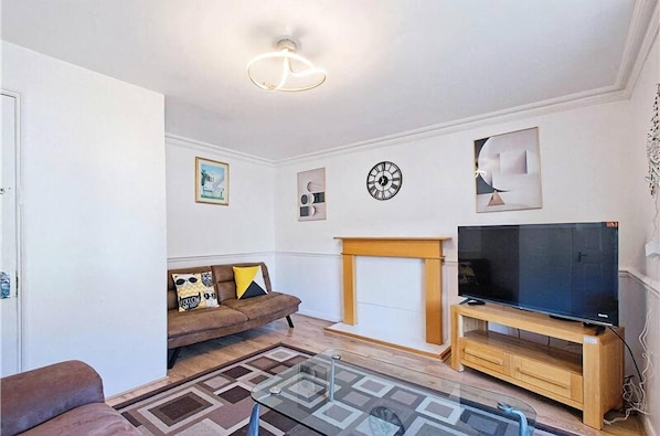 A living room with a sofa, a glass coffee table, a fireplace, a television on a wooden stand, and a clock on the wall. - Apartment - Sleeps 5 - Private Garden (London)
