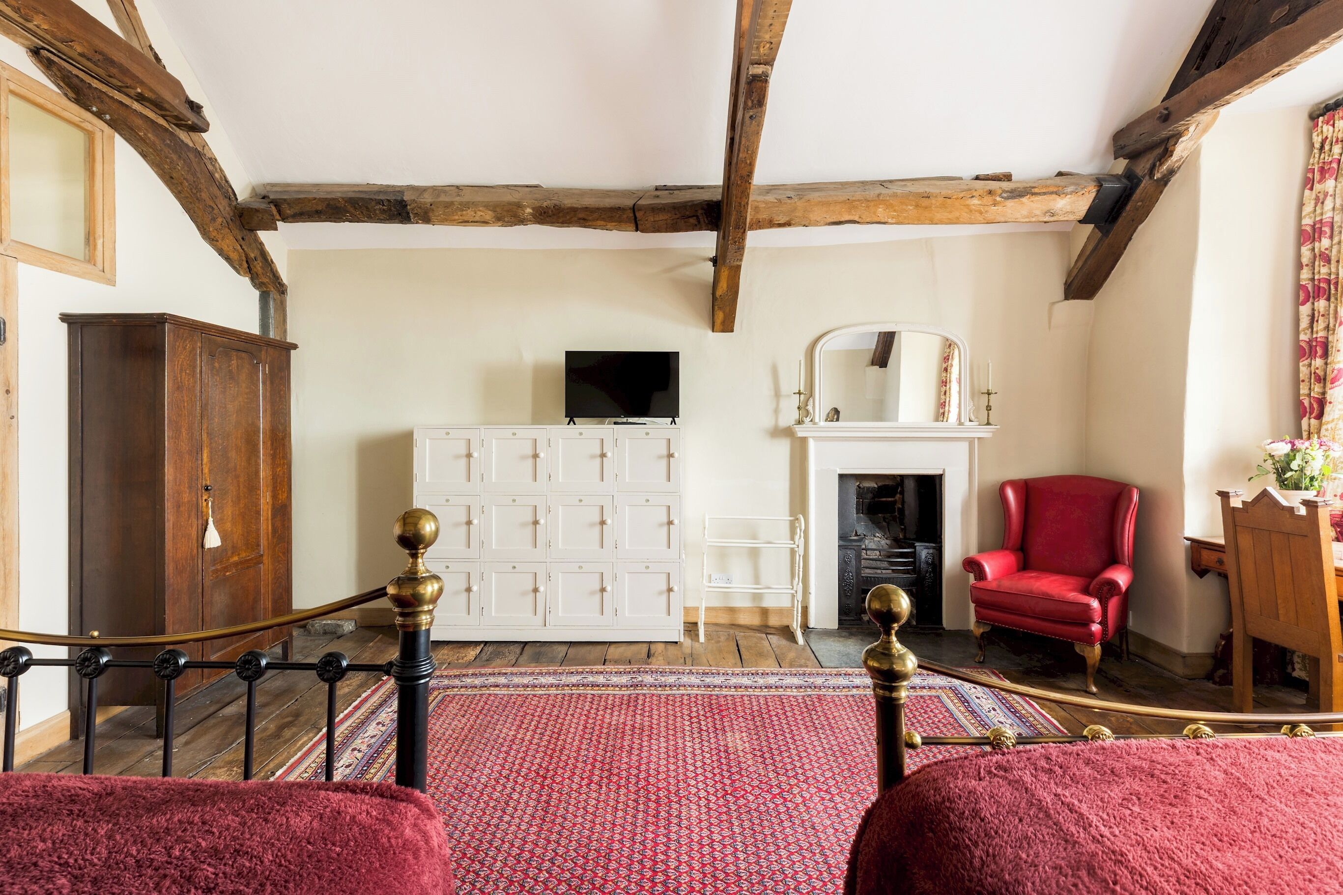 A room with a wooden ceiling, a bed with a red blanket, a red armchair, a white cabinet, and a fireplace.