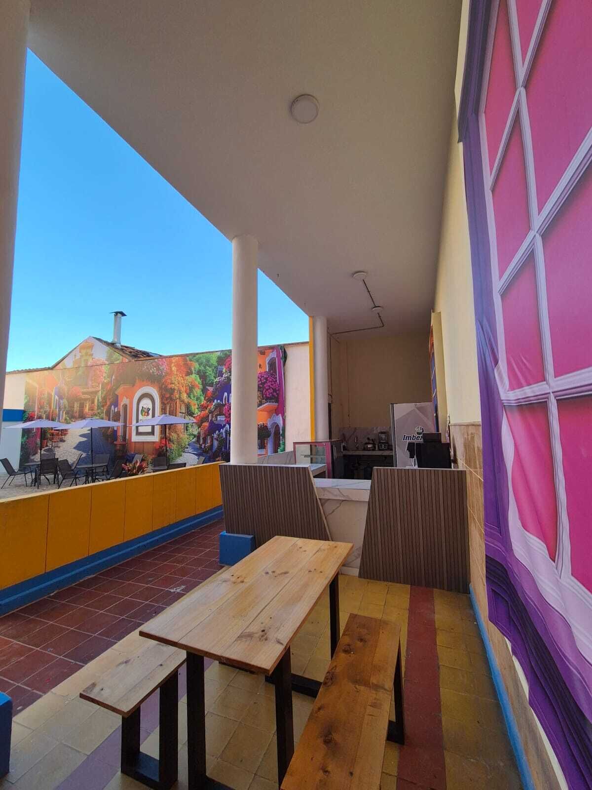 Un café en plein air avec des tables et des chaises en bois, une fresque murale et une vue sur un bâtiment doté d’une cheminée.