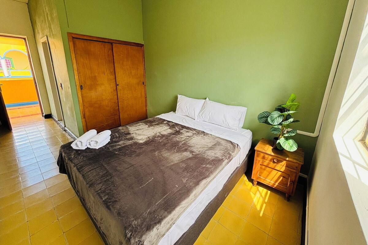 A room with a bed, a wooden wardrobe, a nightstand with a plant, and a tiled floor.