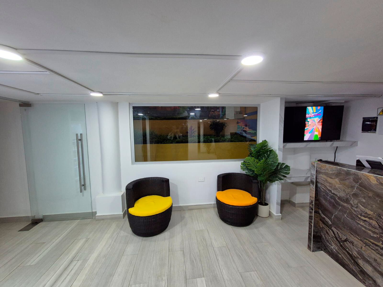 Un área de recepción con dos sillones de mimbre negro con cojines amarillos, una planta en maceta, un televisor en la pared y un mostrador de recepción de mármol.