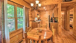 Ein Holzblock-Innenraum mit Esstisch, Stühlen, einer Chandelieber und Blick auf den Wald durch große Fenster.
