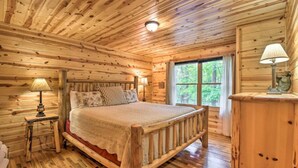 A wooden cabin bedroom with a bed, two bedside lamps, and a window with a view of trees.