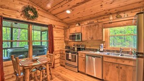 A wooden cabin kitchen with a dining table, stainless steel appliances, and large windows overlooking a forest.