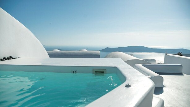 Uma piscina infinita branca moderna com águas azuis cristalinas, rodeada por estruturas arquitetônicas brancas, oferecendo vista para o mar e montanhas ao longe.