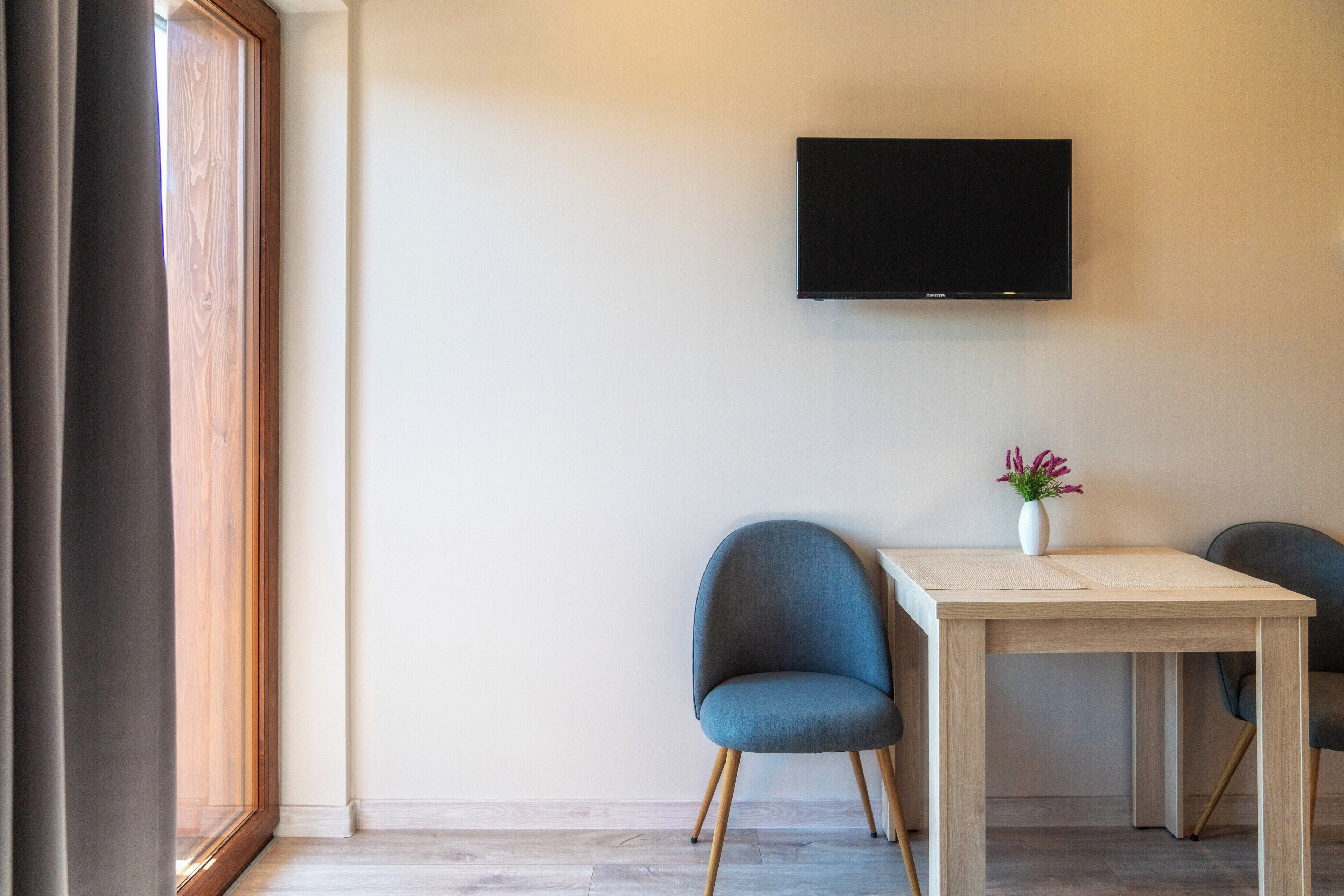 A modern room with a flat-screen TV mounted on the wall, a wooden table, two blue chairs, and a vase with flowers.