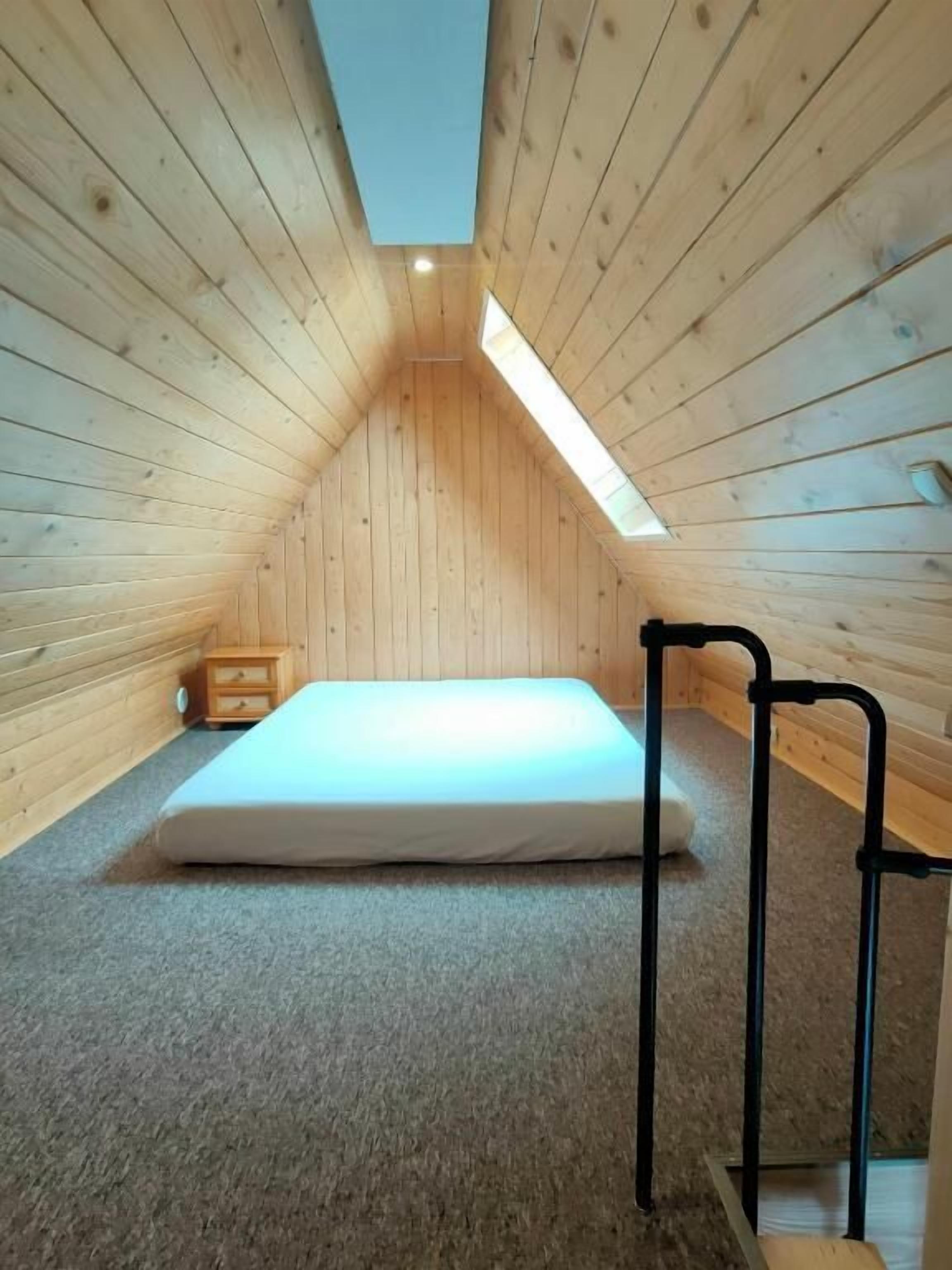 A small, sloped-roofed room with a single mattress on the floor, a wooden bench, and a skylight.