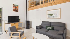 Ein modernes Wohnzimmer mit grauem Sofa, einem kleinen weißen Tisch und einem Holzstuhl. Ein Fernseher steht auf einer Holzkommode, und es gibt einen Kamin sowie einen Balkon mit Holzbalkonen.