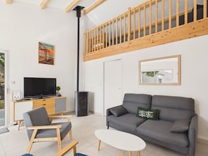A modern living room with a grey sofa, a small white table, and a wooden chair. There's a TV on a wooden cabinet, a fireplace, and a balcony with wooden railings. - House for 7 with garden, beach nearby (Notre-Dame-de-Monts)