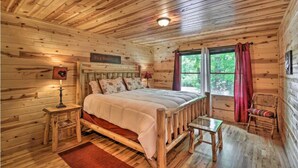 Ein Schlafzimmer einer Holzhütte mit einem Bett, einem Nachttisch, einem Stuhl und einem Fenster mit Vorhängen.