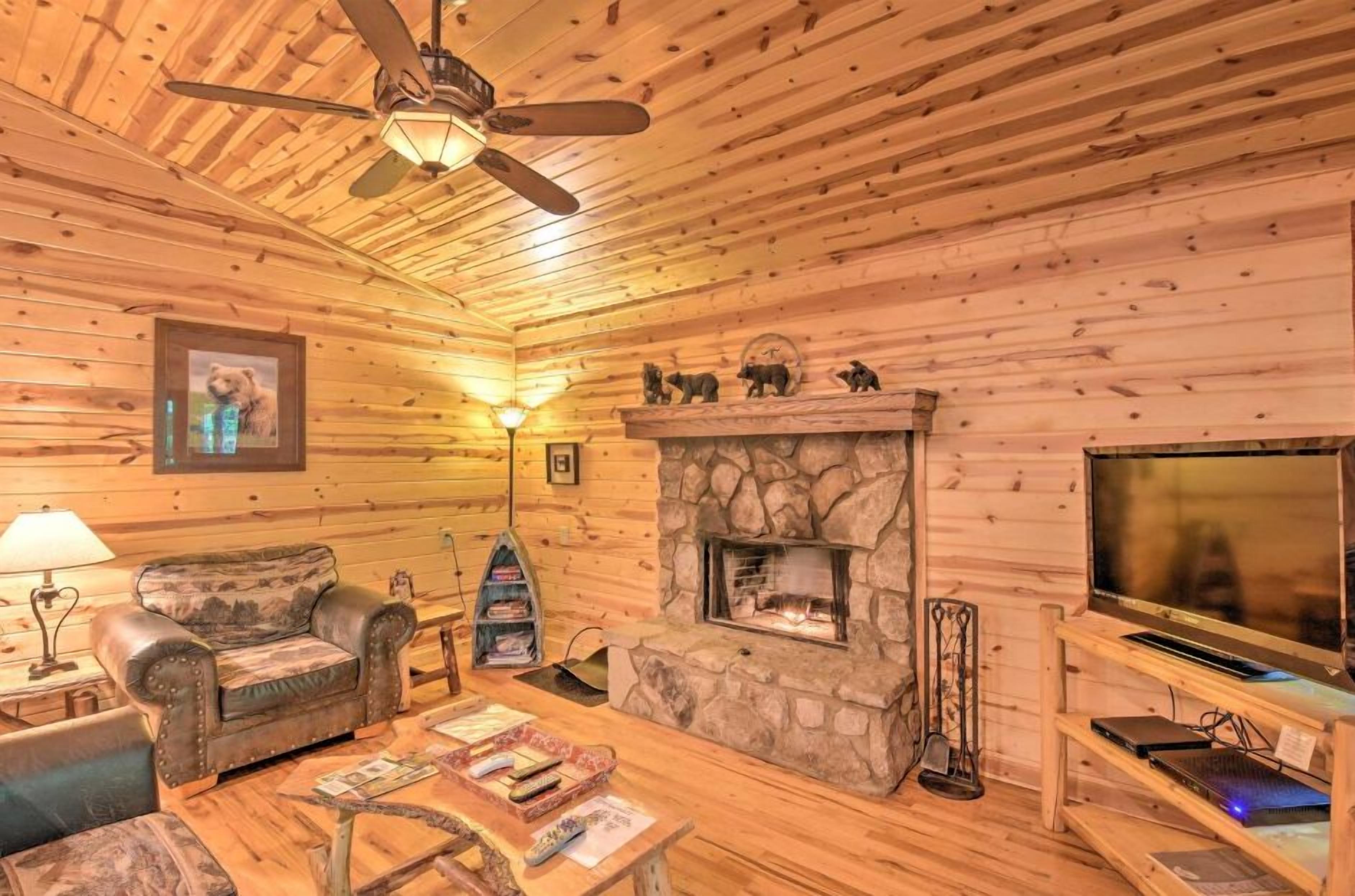 A cozy cabin interior with a stone fireplace, wooden walls, a ceiling fan, and a television.