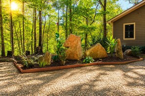 Une maison entourée d’une forêt, avec un chemin de gravier et de gros rochers dans le jardin.