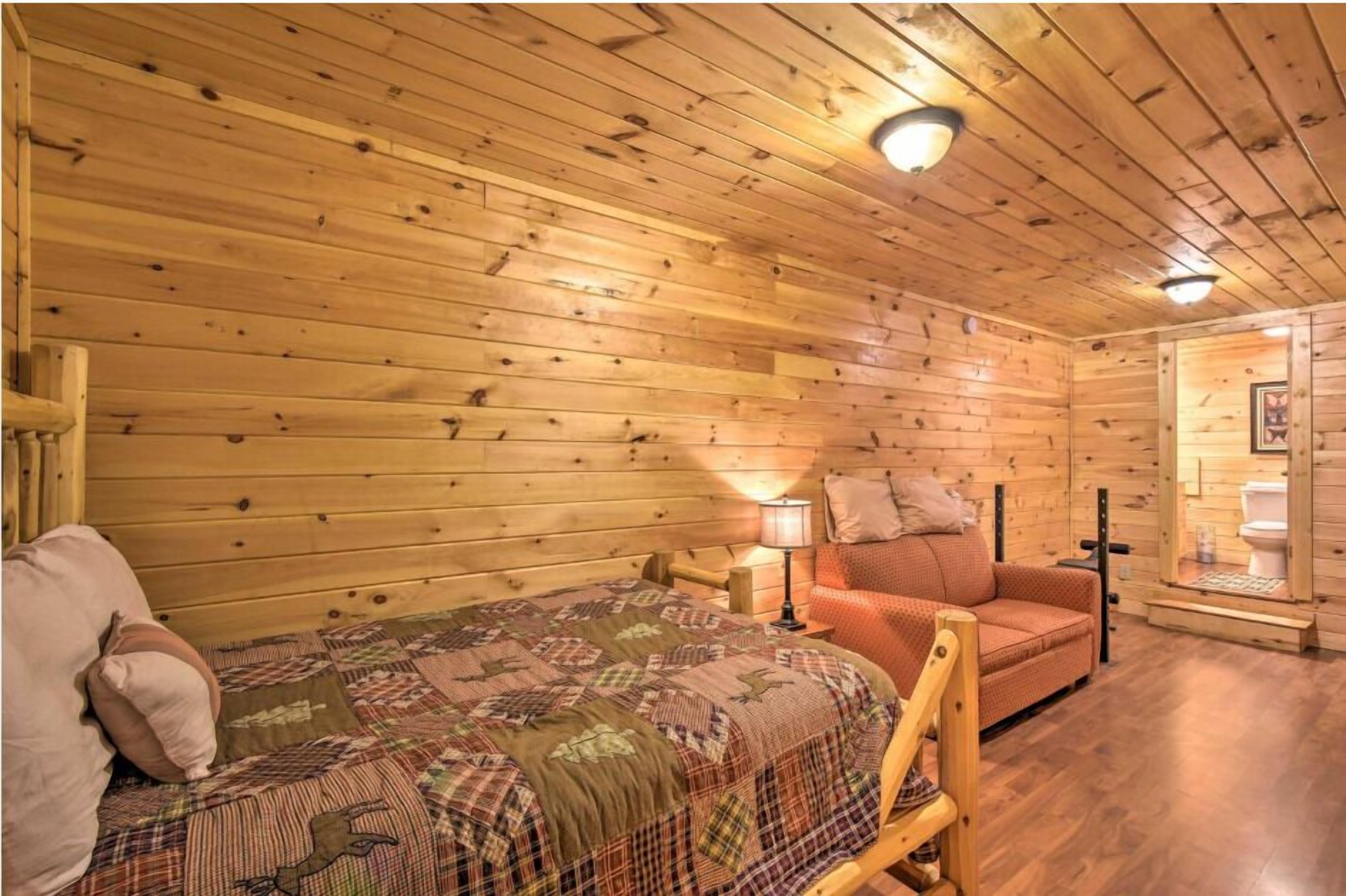 A cozy cabin-style room with wooden walls and a bed with a quilt. There is a small table with a lamp and a sofa with cushions.