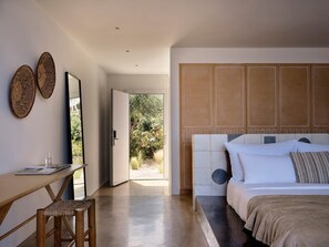 A bedroom with a large bed, a wooden headboard, a desk with a chair, and a view of a garden through an open door. - Veloria Suite, Divine Sea Views, Infinity Pool, Oia (Santorini)