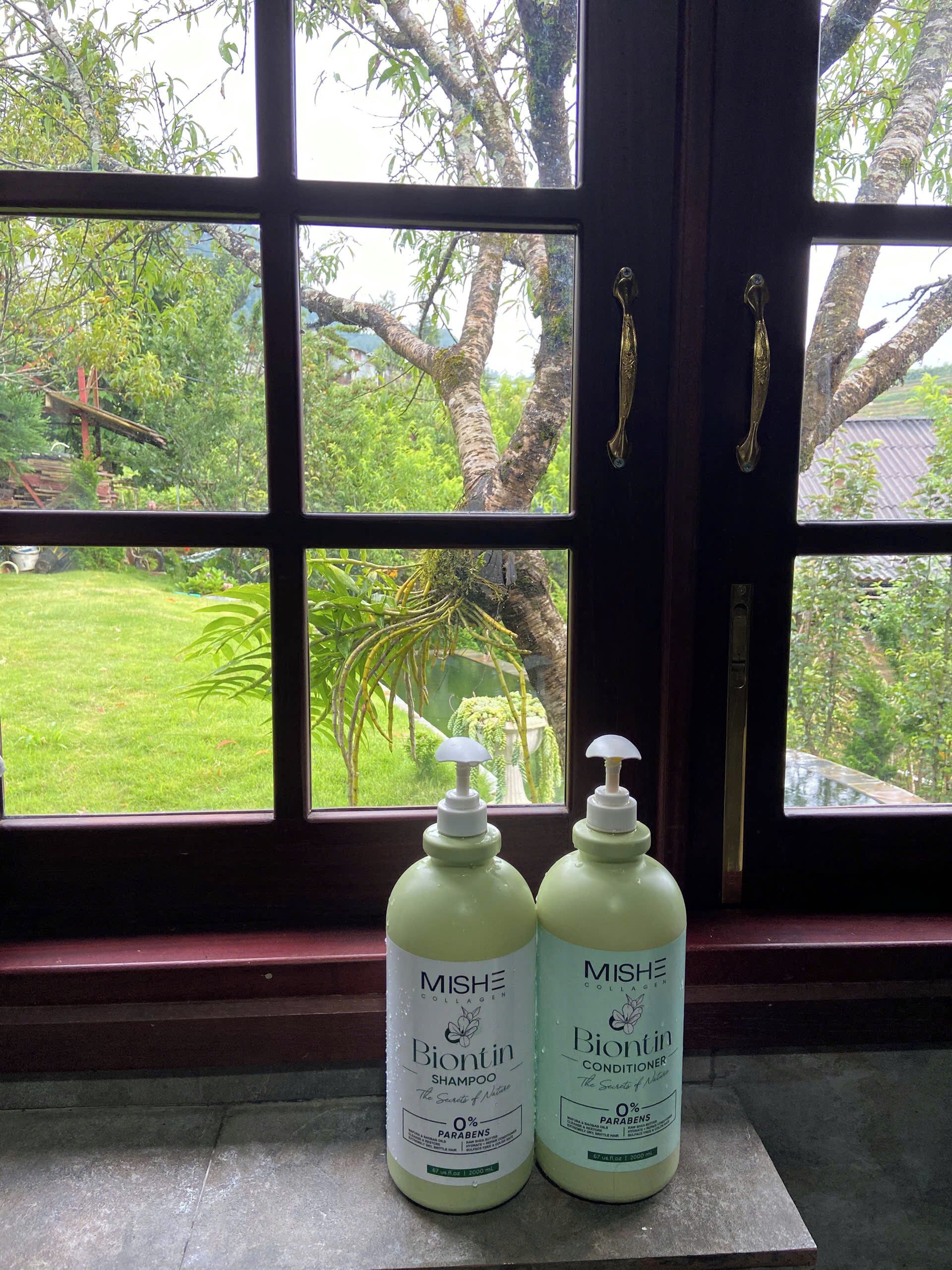 Two bottles of Mische Biontin shampoo on a windowsill with a view of a garden and trees outside.