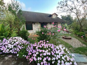 A stone house with a sloped roof, surrounded by a garden with pink and white flowers.