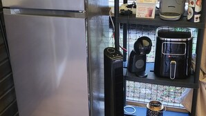 Une cuisine de taille réduite, dotée d’un réfrigérateur en acier inoxydable, d’un ventilateur noir et d’une machine à café posée sur une étagère.