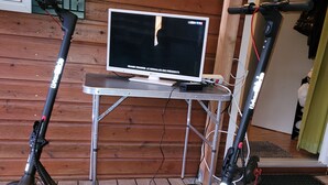 Deux trottinettes électriques sont garées sur une terrasse en bois, et on aperçoit une télévision posée sur une table en métal à l’arrière-plan.