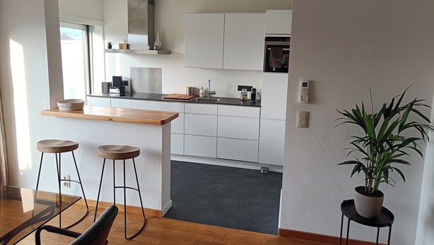 A modern kitchen with white cabinets, a wooden countertop, and a built-in oven. There are stools at a dining table and a potted plant on a side table. - Stylish apartment with AC and sweeping views over historic Leuven (Leuven)