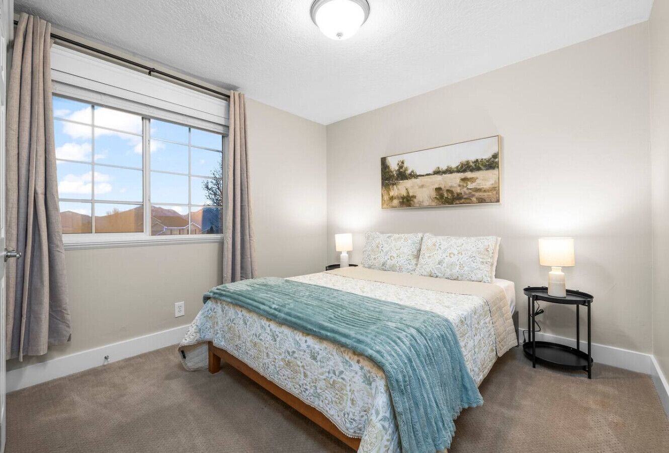 Une chambre à coucher comprenant un lit, deux lampes de chevet, une fenêtre avec des rideaux, un tableau accroché au mur et un sol recouvert d’une moquette.