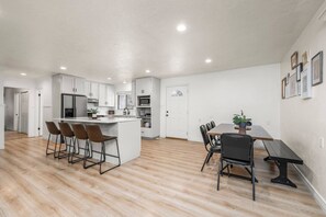 A modern kitchen with white cabinets, a central island with bar stools, a dining area with a wooden table and chairs, and a hallway leading to another room.
