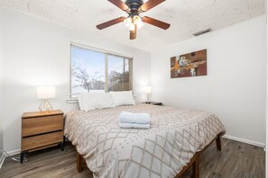 A bedroom with a bed, two bedside tables, a ceiling fan, a window with blinds, and a painting on the wall.