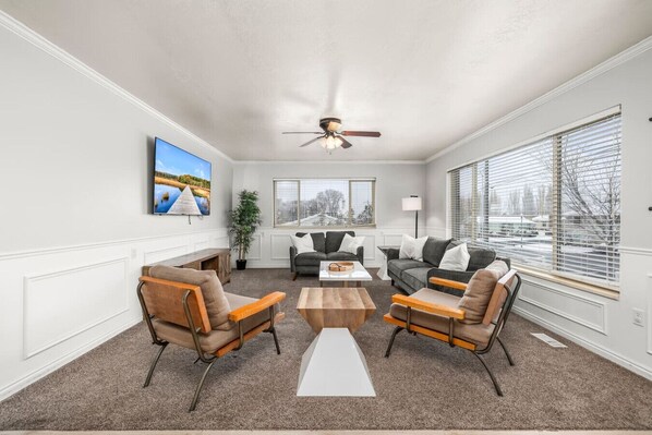 A spacious living room with a ceiling fan, large windows with blinds, a flat-screen TV, and two sofas.
