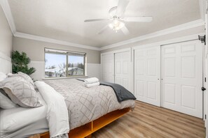 A bedroom with a bed, a ceiling fan, a window with a view, and a set of white doors.