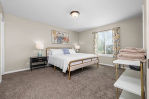 A bedroom with a bed, bedside tables, a window with curtains, a painting on the wall, and a stack of towels on a chair. - Cozy Orem House | Playground | Game Rm | Bunk Rm (Orem)