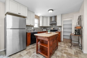 A modern kitchen with white cabinets, a stainless steel refrigerator, a wooden island, and a stone backsplash. - Cozy Orem House | Playground | Game Rm | Bunk Rm (Orem)