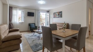 A living room with a sofa, a dining table with chairs, a TV mounted on the wall, and a painting. - Apartment Seralya, Spacious Two Bedroom Apartment, Athens (Athens)