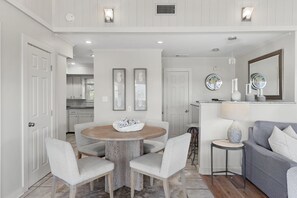 A modern kitchen with white cabinetry, a dining area with a round wooden table and four chairs, and a gray sofa in a living area. - Coastal Bungalow Retreat,  Bikes & Spacious Sun Deck – Minutes to the Beach (Inlet Beach)
