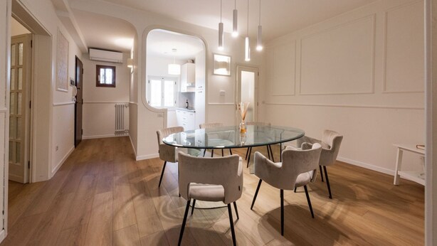 Et moderne spiseområde med et glasbord og stole, et køkken i baggrunden og en gang, der fører til andre værelser.