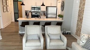 Un salon moderne avec une table basse en bois, deux fauteuils, un canapé et une cuisine en arrière-plan.
