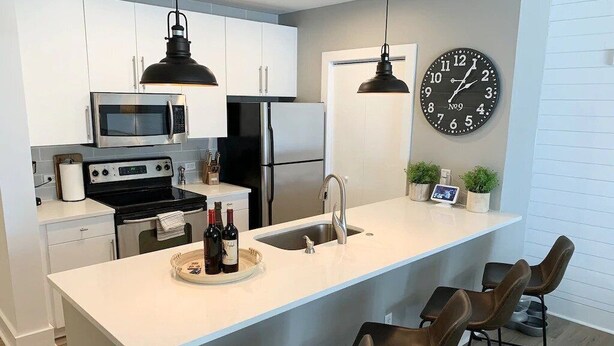 Une cuisine moderne avec des meubles blancs, des appareils en acier inoxydable, un grand îlot central doté de tabourets et une horloge murale.
