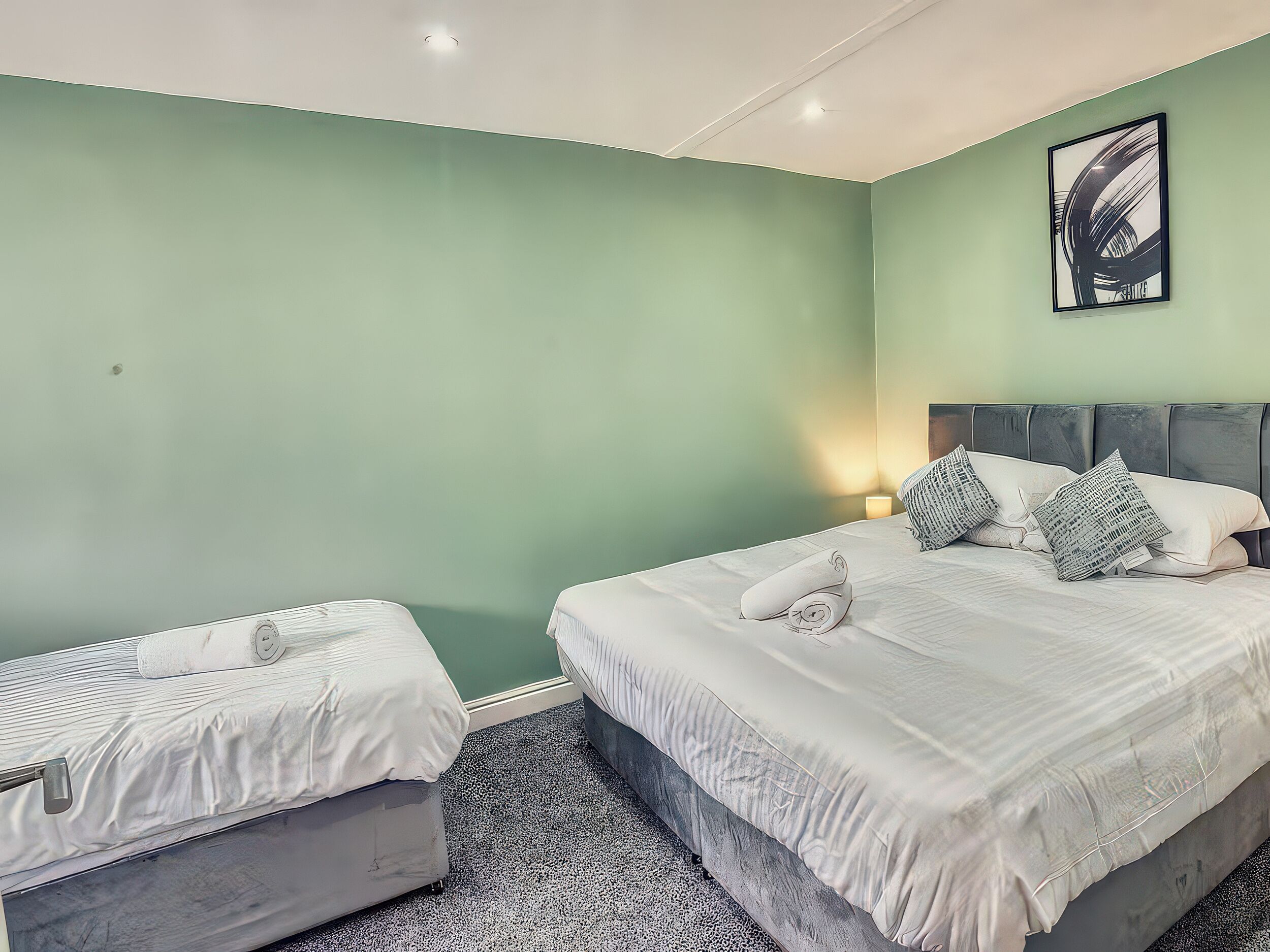 Ein Hotelzimmer mit zwei Betten, einer grünen Wand, einem gerahmten Kunstwerk und einem kleinen Nachttisch mit Lampe.